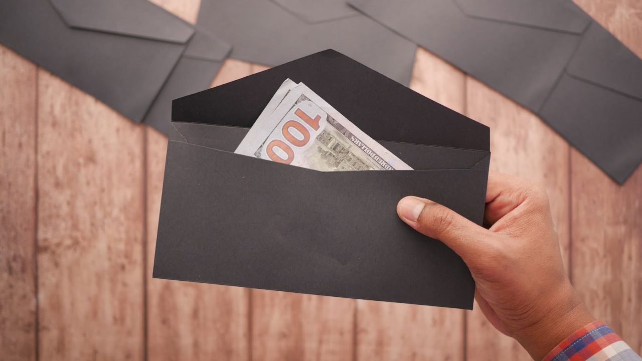 A hand holds a black envelope containing a 100 dollar bill on a wooden surface.